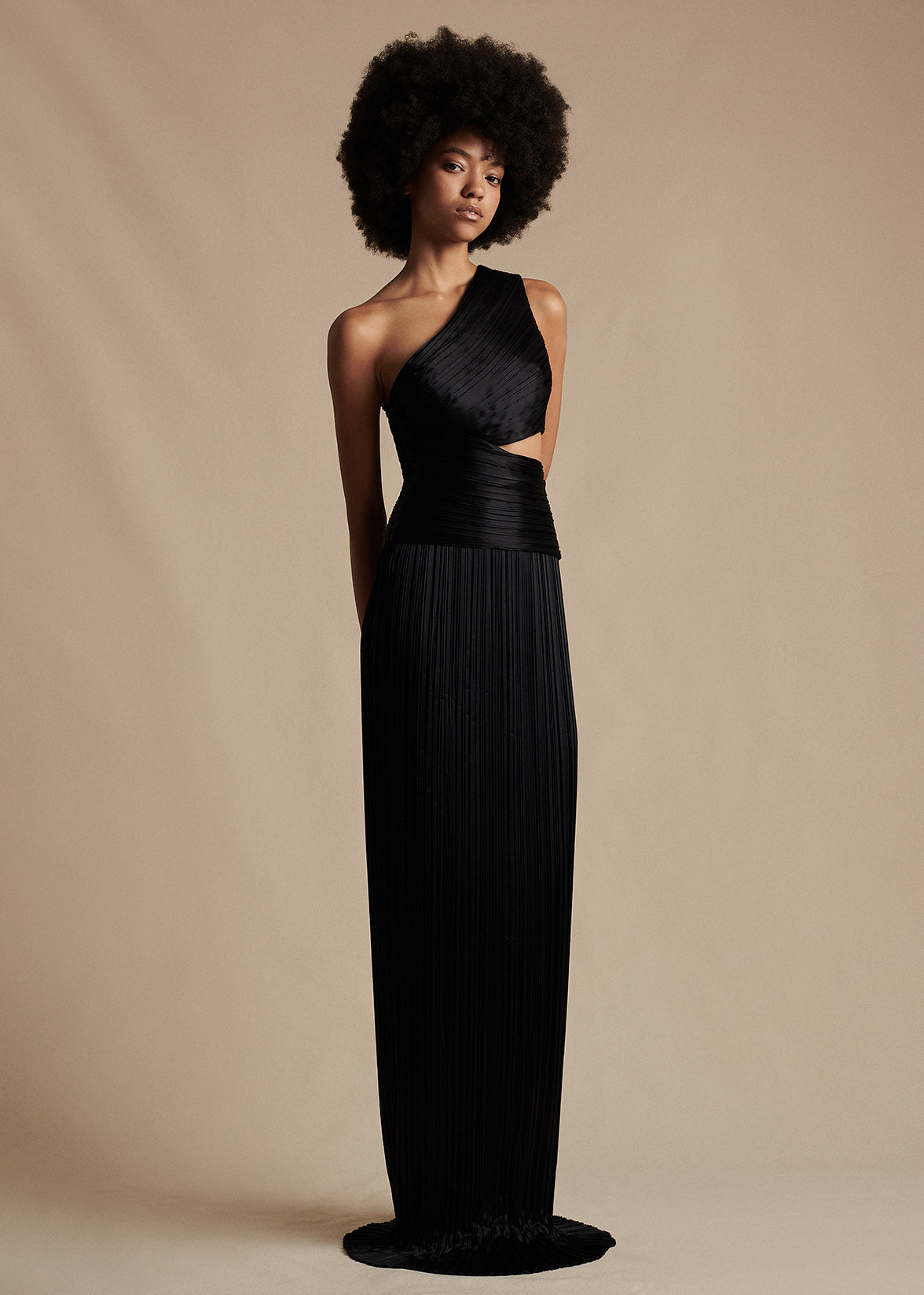 Model is wearing the one-shoulder Delphos Dress in Charmeuse. It is a black, floor-length dress with a cut-out on the wearer's left side.