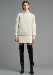 A model wearing an ivory knit turtleneck and matching mini skirt and tall black boots.